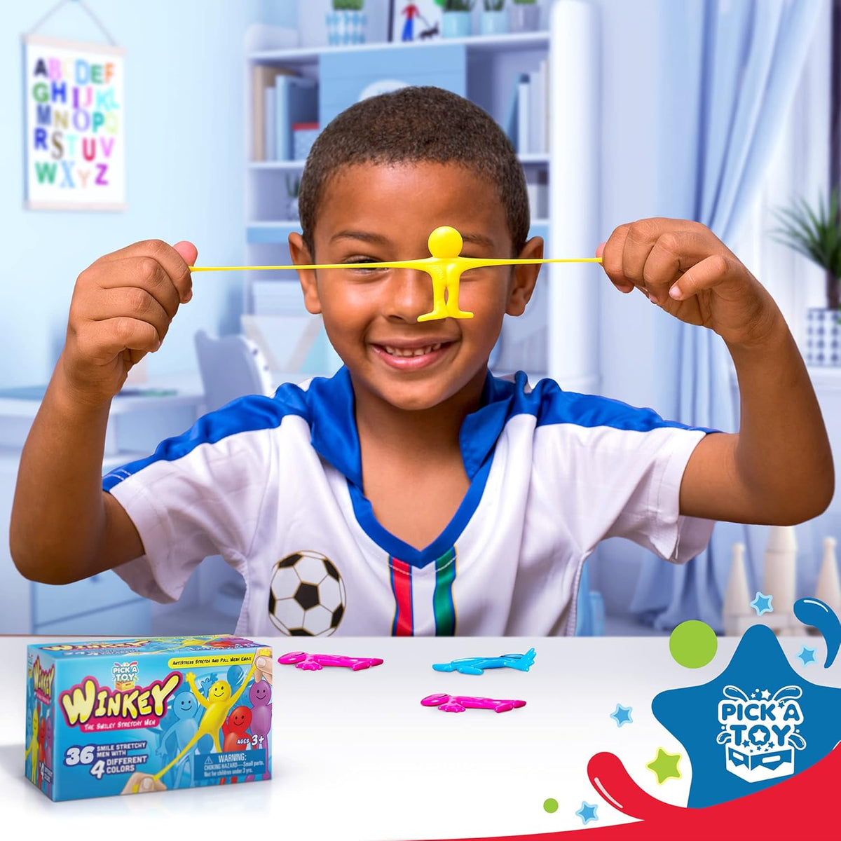 Smiling boy stretching a yellow stretchy man toy across his face at a desk, with the box and more figures nearby